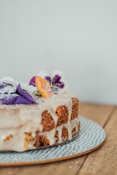 A close-up image of a two-layer cake with white icing, decorated with purple and orange edible flowers and a sprinkle of powdered sugar, placed on a textured, dotted plate. The background is a neutral, plain wall and a wooden surface beneath the plate.