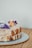 A close-up image of a two-layer cake with white icing, decorated with purple and orange edible flowers and a sprinkle of powdered sugar, placed on a textured, dotted plate. The background is a neutral, plain wall and a wooden surface beneath the plate.