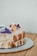 A close-up image of a two-layer cake with white icing, decorated with purple and orange edible flowers and a sprinkle of powdered sugar, placed on a textured, dotted plate. The background is a neutral, plain wall and a wooden surface beneath the plate.