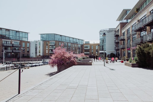 A modern urban area with several multi-story apartment buildings featuring large glass windows and brick facades. The foreground includes a paved plaza with planted trees and shrubs. Outdoor seating areas with tables and chairs are visible along the building exteriors. A few people are walking along the pathway.