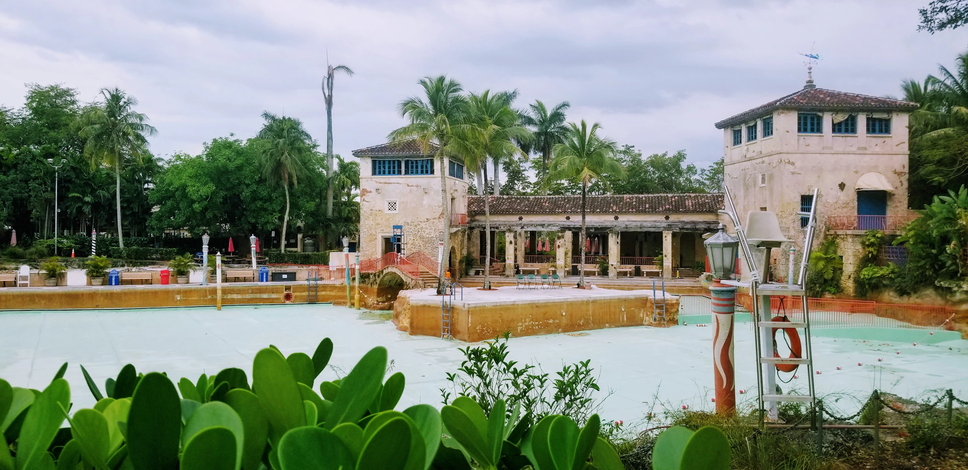 A sepia-toned photo of a beautifully maintained vintage-style swimming pool surrounded by classic wrought iron furniture and lush greenery.