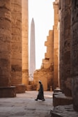 Ancient stone pillars covered with hieroglyphics surround a narrow pathway, where a person wearing traditional clothing walks. In the background, an obelisk stands tall against the bright sky, blending with the warm tones of the architecture.