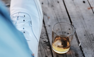 A white sneaker rests on a gray wooden surface next to a small glass containing a light amber liquid.