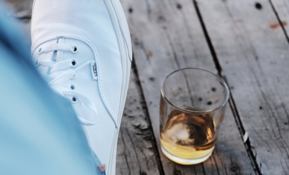 A white sneaker rests on a gray wooden surface next to a small glass containing a light amber liquid.
