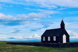 A black wooden church stands alone on a green grassy field under a vast, partly cloudy blue sky. The building features a white door and three windows, surrounded by a low stone wall. The landscape appears open and serene, with a distant horizon visible.