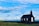 A black wooden church stands alone on a green grassy field under a vast, partly cloudy blue sky. The building features a white door and three windows, surrounded by a low stone wall. The landscape appears open and serene, with a distant horizon visible.
