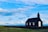A black wooden church stands alone on a green grassy field under a vast, partly cloudy blue sky. The building features a white door and three windows, surrounded by a low stone wall. The landscape appears open and serene, with a distant horizon visible.