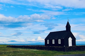 A black wooden church stands alone on a green grassy field under a vast, partly cloudy blue sky. The building features a white door and three windows, surrounded by a low stone wall. The landscape appears open and serene, with a distant horizon visible.