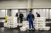 Several people are working in an industrial setting, stacking and organizing white plastic crates. They are wearing work uniforms, and the environment appears to be a storage or processing facility with overhead lighting and large doors in the background.