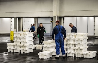 Several people are working in an industrial setting, stacking and organizing white plastic crates. They are wearing work uniforms, and the environment appears to be a storage or processing facility with overhead lighting and large doors in the background.
