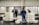 Several people are working in an industrial setting, stacking and organizing white plastic crates. They are wearing work uniforms, and the environment appears to be a storage or processing facility with overhead lighting and large doors in the background.