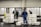 Several people are working in an industrial setting, stacking and organizing white plastic crates. They are wearing work uniforms, and the environment appears to be a storage or processing facility with overhead lighting and large doors in the background.