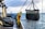A worker in a yellow and red waterproof suit stands on a boat deck, operating machinery to hoist a large net filled with seaweed or marine debris from the ocean. The scene is set against a backdrop of a cloudy sky and calm sea.