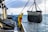 A worker in a yellow and red waterproof suit stands on a boat deck, operating machinery to hoist a large net filled with seaweed or marine debris from the ocean. The scene is set against a backdrop of a cloudy sky and calm sea.