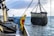 A worker in a yellow and red waterproof suit stands on a boat deck, operating machinery to hoist a large net filled with seaweed or marine debris from the ocean. The scene is set against a backdrop of a cloudy sky and calm sea.
