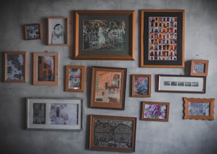 A collection of various framed artworks hangs on a textured wall. The frames vary in size and style, showcasing a mix of paintings, illustrations, and photographs. These include depictions of urban architecture, a garden scene, abstract art, and floral patterns. The arrangement is eclectic, suggesting a personal or curated collection.