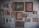 A collection of various framed artworks hangs on a textured wall. The frames vary in size and style, showcasing a mix of paintings, illustrations, and photographs. These include depictions of urban architecture, a garden scene, abstract art, and floral patterns. The arrangement is eclectic, suggesting a personal or curated collection.