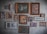 A collection of various framed artworks hangs on a textured wall. The frames vary in size and style, showcasing a mix of paintings, illustrations, and photographs. These include depictions of urban architecture, a garden scene, abstract art, and floral patterns. The arrangement is eclectic, suggesting a personal or curated collection.