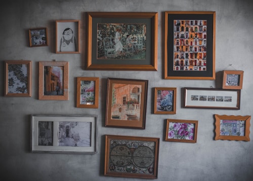 A collection of various framed artworks hangs on a textured wall. The frames vary in size and style, showcasing a mix of paintings, illustrations, and photographs. These include depictions of urban architecture, a garden scene, abstract art, and floral patterns. The arrangement is eclectic, suggesting a personal or curated collection.