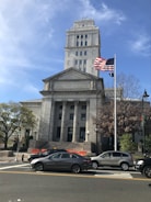 A neoclassical courthouse building stands prominently with tall columns and a square tower. In front, an American flag waves on a flagpole. Several cars are parked or waiting on the road, and pedestrians can be seen near traffic signals and trees. The architecture is grand and historic, reflecting governmental importance.