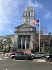 A neoclassical courthouse building stands prominently with tall columns and a square tower. In front, an American flag waves on a flagpole. Several cars are parked or waiting on the road, and pedestrians can be seen near traffic signals and trees. The architecture is grand and historic, reflecting governmental importance.