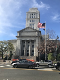 A neoclassical courthouse building stands prominently with tall columns and a square tower. In front, an American flag waves on a flagpole. Several cars are parked or waiting on the road, and pedestrians can be seen near traffic signals and trees. The architecture is grand and historic, reflecting governmental importance.
