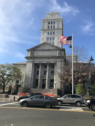 A neoclassical courthouse building stands prominently with tall columns and a square tower. In front, an American flag waves on a flagpole. Several cars are parked or waiting on the road, and pedestrians can be seen near traffic signals and trees. The architecture is grand and historic, reflecting governmental importance.