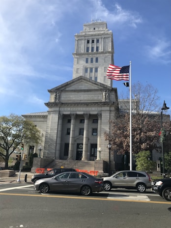 A neoclassical courthouse building stands prominently with tall columns and a square tower. In front, an American flag waves on a flagpole. Several cars are parked or waiting on the road, and pedestrians can be seen near traffic signals and trees. The architecture is grand and historic, reflecting governmental importance.