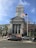 A neoclassical courthouse building stands prominently with tall columns and a square tower. In front, an American flag waves on a flagpole. Several cars are parked or waiting on the road, and pedestrians can be seen near traffic signals and trees. The architecture is grand and historic, reflecting governmental importance.