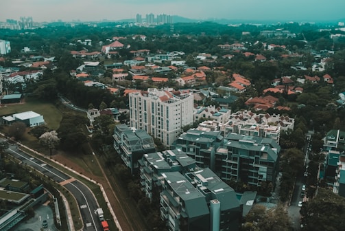 Aerial view of a residential area with a mix of high-rise buildings and lower residential houses. The landscape includes a blend of densely packed structures and green, tree-covered areas. Buildings have varied architectural styles, surrounded by winding roads and scattered vegetation.