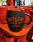 A black ceramic mug with red interior and red text reading 'RUTGERS UNIVERSITY' placed on a wooden shelf. Several other red mugs are visible in the background. The setting appears to be a store or shop with blurred merchandise visible in the background.