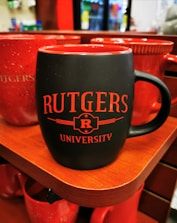 A black ceramic mug with red interior and red text reading 'RUTGERS UNIVERSITY' placed on a wooden shelf. Several other red mugs are visible in the background. The setting appears to be a store or shop with blurred merchandise visible in the background.