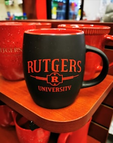 A black ceramic mug with red interior and red text reading 'RUTGERS UNIVERSITY' placed on a wooden shelf. Several other red mugs are visible in the background. The setting appears to be a store or shop with blurred merchandise visible in the background.