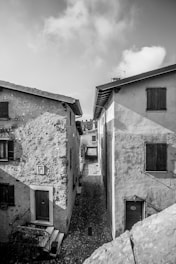 A monochrome image capturing a narrow, cobblestone alley lined with weathered stone buildings and soft shadows.