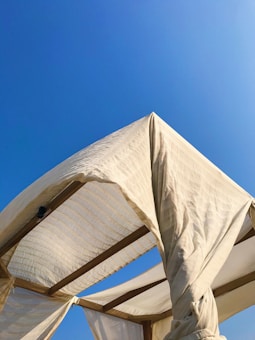 A wooden structure with draped white fabric creates a shaded area under a clear blue sky. The fabric twists around the beams, casting shadows and creating a soft, flowing effect.