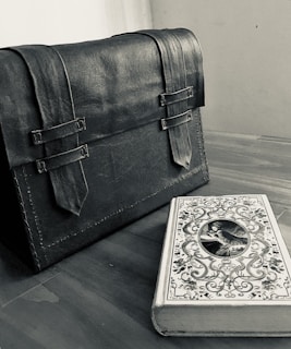 A leather satchel with two buckled straps sits on a wooden surface next to a decorative book with intricate designs and an oval portrait on the cover.