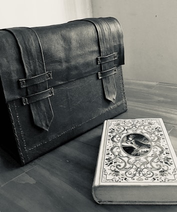 A leather satchel with two buckled straps sits on a wooden surface next to a decorative book with intricate designs and an oval portrait on the cover.