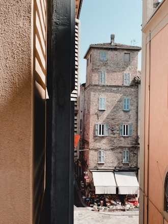A narrow alleyway framed by tall buildings leads to a small open market area. The historical building in view has brick walls and several windows with closed shutters. A variety of kitchen and market goods are displayed under white canopies at street level.