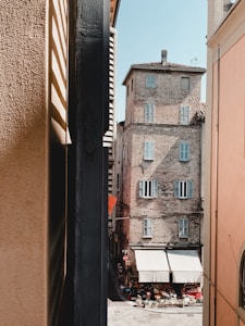 A narrow alleyway framed by tall buildings leads to a small open market area. The historical building in view has brick walls and several windows with closed shutters. A variety of kitchen and market goods are displayed under white canopies at street level.