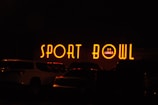 The image depicts a brightly lit neon sign that reads 'SPORT BOWL' with an additional sign indicating '24 LANES'. The scene appears to be taken at night, with several vehicles parked in front of the establishment.