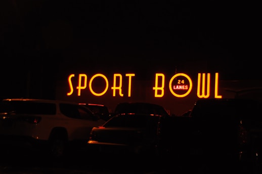 The image depicts a brightly lit neon sign that reads 'SPORT BOWL' with an additional sign indicating '24 LANES'. The scene appears to be taken at night, with several vehicles parked in front of the establishment.
