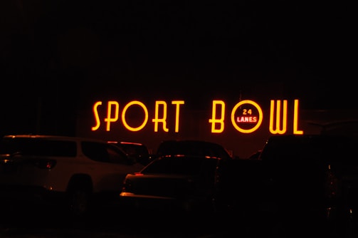 The image depicts a brightly lit neon sign that reads 'SPORT BOWL' with an additional sign indicating '24 LANES'. The scene appears to be taken at night, with several vehicles parked in front of the establishment.