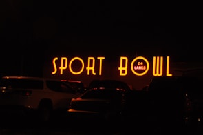 The image depicts a brightly lit neon sign that reads 'SPORT BOWL' with an additional sign indicating '24 LANES'. The scene appears to be taken at night, with several vehicles parked in front of the establishment.