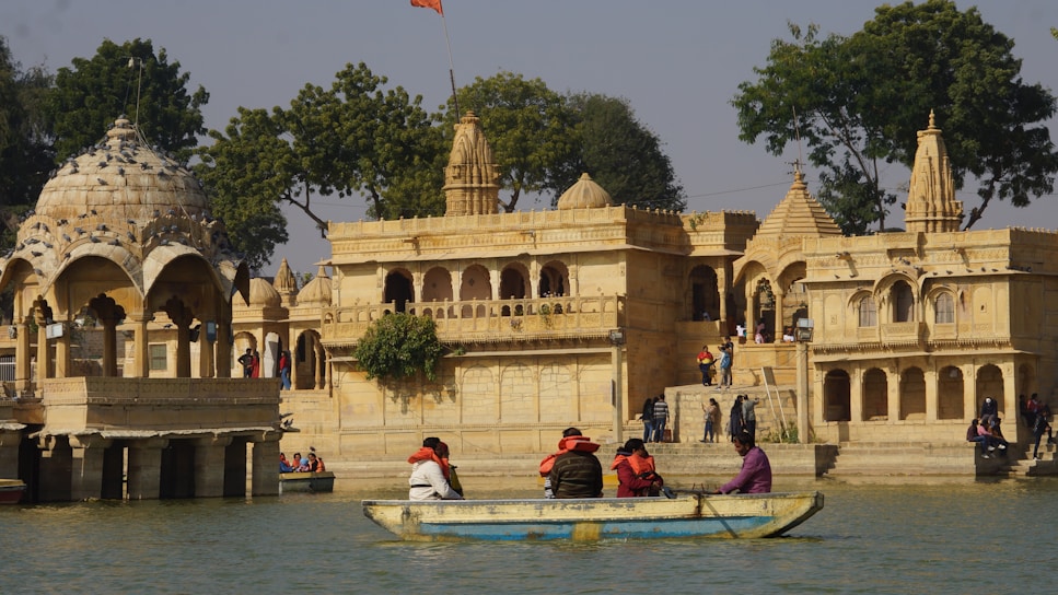 A historical architecture featuring ornate structures with domes and intricate carvings is set near a water body. A small group of people wearing colorful clothing is sitting in a boat on the water, while others are walking around the architectural site. The scene is surrounded by lush greenery and a few trees.