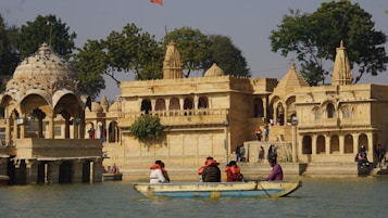 A historical architecture featuring ornate structures with domes and intricate carvings is set near a water body. A small group of people wearing colorful clothing is sitting in a boat on the water, while others are walking around the architectural site. The scene is surrounded by lush greenery and a few trees.