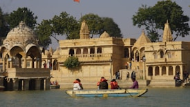 A historical architecture featuring ornate structures with domes and intricate carvings is set near a water body. A small group of people wearing colorful clothing is sitting in a boat on the water, while others are walking around the architectural site. The scene is surrounded by lush greenery and a few trees.
