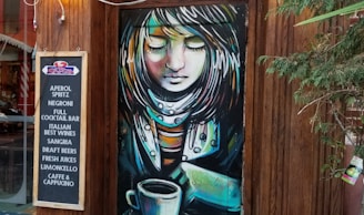 A vibrant mural depicting a stylized female figure with closed eyes is painted on a wooden door. The figure has colorful hair and a serene expression. Next to the mural is a chalkboard menu listing drinks such as Aperol Spritz, Negroni, and various Italian beverages. The setting includes wooden elements and some greenery.