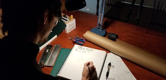 A person is drawing in a sketchbook at a wooden table. The scene is illuminated by a desk lamp, creating a focused workspace. Various art supplies are scattered around, including pencils, a compass, and a partially unrolled sheet of brown paper.