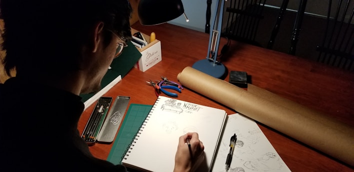 A person is drawing in a sketchbook at a wooden table. The scene is illuminated by a desk lamp, creating a focused workspace. Various art supplies are scattered around, including pencils, a compass, and a partially unrolled sheet of brown paper.