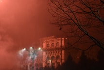 A dramatic scene features an illuminated building shrouded in red mist. Bare tree branches are silhouetted against the sky, adding a sense of eerie isolation. Streetlights cast a smoky glow over the area, enhancing the mysterious atmosphere.